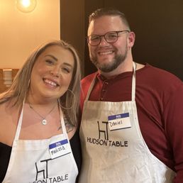 A woman with shoulder-length blonde hair and a man with glasses and a beard are standing close together, smiling at the camera. They are wearing aprons with the Hudson Table logo and name tags reading 'Melissa' and 'Daniel.' The background includes warm lighting and a small plant on a shelf, indicating an indoor environment, likely at a culinary or event space.