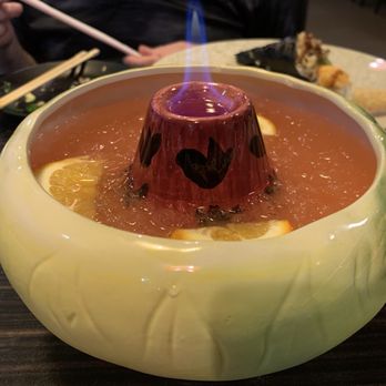 Osaka - A large, decorative ceramic bowl filled with fruit punch containing lemon slices and ice, with an ice sculpture in the center. The bowl is placed on a dark wooden table in a restaurant setting. In the background, a person is holding chopsticks, and a plate with sushi is visible. - Asian Fusion Near Me - Bloomfield, Connecticut