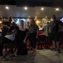 Six people are standing together in a bar with a dark interior, bar counter, and patrons seated in the background. The lighting is low.