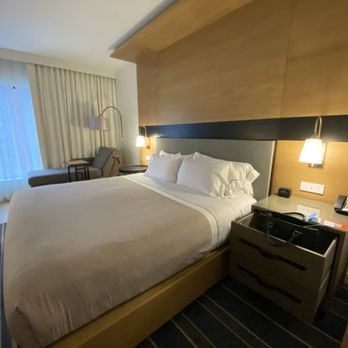 Canopy by Hilton Ithaca Downtown - Nov, 2024. - The image shows a hotel room with a large bed covered in white linens and pillows. To the right of the bed is a nightstand holding a phone, clock, and some papers. The headboard extends across the wall, with wall-mounted lamps on each side. In the background, there is a seating area with a chair, a small table, and a floor lamp near a window with curtains. The room has a modern, clean design with neutral colors and wooden accents. - Hotels Near Me - Ithaca, New York