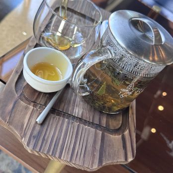Book Bar - Tray with tea service - A glass teapot containing loose leaf tea, a glass cup with tea, and a small white dish with tea are placed on a wooden tray. The tray is on a table, with chairs and a patterned rug visible in the background. The setting appears to be a cafe or tea shop. - Sandwiches Near Me - Mokena, Illinois