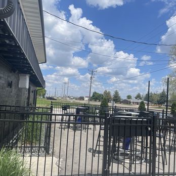 Sunset Lounge - Outdoor Patio - An outdoor patio area with tables and chairs, enclosed by a black metal fence, adjacent to a building with a black brick exterior. - Burgers Near Me - Norwalk, Iowa