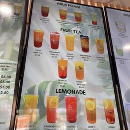 Menu - The menu board features sections for milk foam, fruit tea, and lemonade drinks, each with images, names, and prices. Milk foam options include peach, green tea, jasmine tea, strawberry matcha, and coconut juice. Fruit teas include peach, strawberry, mango, wintermelon, grapefruit, tropical, and kumquat flavors. Lemonades include passionfruit, strawberry, honey, and lychee, all priced at $5.29. The board is decorated with green leaf graphics, and the drinks are visually represented with colorful images.