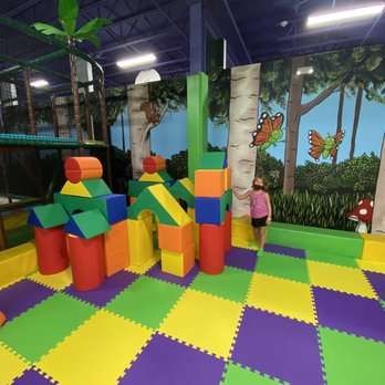 Fireflies Fun Zone - Building blocks - A girl in a pink shirt and black shorts stands in an indoor play zone with foam blocks, a mural of trees and butterflies, and a padded floor. - Arcades Near Me - Chesterton, Indiana