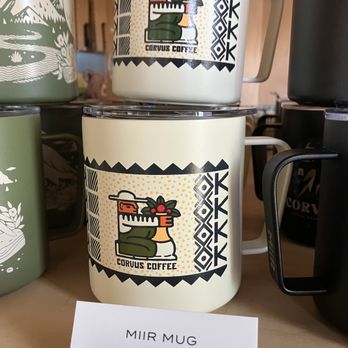 Corvus Coffee Roasters - Two Corvus Coffee mugs featuring a stylized graphic of a person holding a plant are shown, with one mug stacked on top of the other, surrounded by other mugs. - Coffee & Tea Near Me - Arvada, Colorado