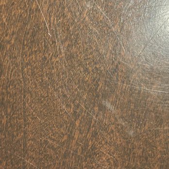 Sweeps - Wood table  and couch - The image depicts a close-up view of a wooden table surface with a dark brown finish. The surface has visible scratches and marks, indicating wear and usage over time. The lighting highlights the texture and imperfections on the wood. - Landscaping Near Me