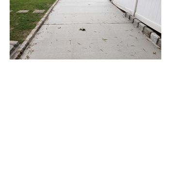 Masone Masonry - Driveway - Concrete driveway with visible cracks and scattered leaves and small debris. It is bordered by a white fence on the right side and a grassy area with plants on the left. The driveway leads to a street in the background, with a curb and sidewalk visible. The scene appears to be outdoors in a residential area. - Masonry/Concrete Near Me