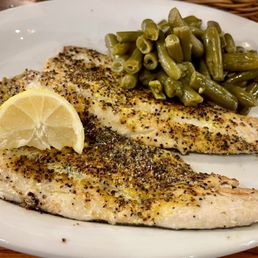Rainbow Trout - A white plate holds a serving of lemon pepper grilled rainbow trout, seasoned with black pepper and herbs, accompanied by cooked green beans. A lemon wedge is placed on top of the fish for added flavor. The dish is presented on a white ceramic plate, with a basket visible in the background.
