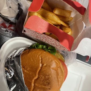 Fat Shack - burger and fries - A cheeseburger with lettuce, tomato, and cheese is partially wrapped in foil, placed on a tray. Next to it, there is a red and white cardboard container filled with French fries. The background includes various food packaging and a plastic bag, indicating a fast-food meal setting. - Sandwiches Near Me - Eugene, Oregon