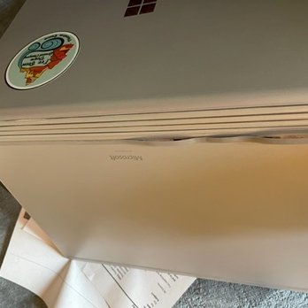 Dotfoil Computer Services - You can see it's a stick on rubber piece... - A silver MacBook laptop with stickers on the lid, resting on a surface with papers underneath, viewed from an angle. - IT Services & Computer Repair Near Me - Santa Fe, New Mexico