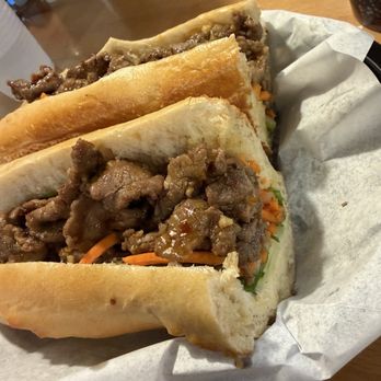 Wrap N Roll - Lemongrass steak bahn mi - Three beef banh mi sandwiches with sliced bread, cooked beef, and vegetables such as carrots and greens are placed on a paper-lined tray. The sandwiches are arranged in a row on a wooden table, with some condiments and drinks visible in the background. - Vietnamese Near Me - Albuquerque, New Mexico