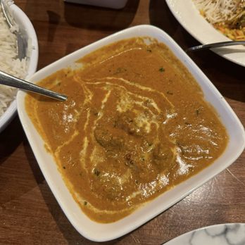 The Mint Leaf - Akhroti Gosht - A square white bowl holds Indian curry with a thick, orange-brown sauce and visible meatballs, garnished with a drizzle of cream or yogurt. The bowl is on a wooden table, with portions of rice dishes in white bowls visible on either side. The setting suggests a restaurant meal featuring traditional Indian cuisine. - Indian Near Me - Cockeysville, Maryland