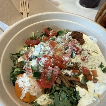 CAVA - Braised lamb with spicy meatballs plus veggies - A disposable bowl holds braised lamb, spicy meatballs, chopped tomatoes, greens, and creamy sauce, with a fork and napkin nearby. - Mediterranean Near Me - Topeka, Kansas