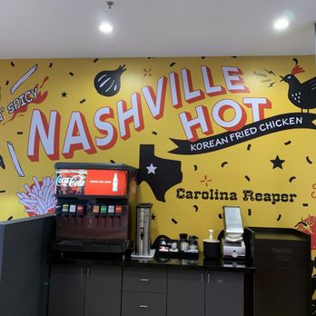 Mighty Chick Sando - The image shows a restaurant interior with a bright yellow wall decorated with a mural advertising Nashville Hot Korean Fried Chicken. The mural includes large text, illustrations of a chicken, Texas shape, and condiments, along with phrases like 'Sweet n' Spicy' and 'Carolina Reaper.' A beverage station with a Coca-Cola dispenser and other drink supplies is positioned below the mural. The ceiling has recessed lighting, and part of a table with a red booth is visible on the left side. - Chicken Wings Near Me - Bedford, Texas