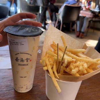 TP TEA - Jasmine Green Milk Tea with Lychee Jelly and Sour Cream & Onion fries - A hand is holding a cup of milk tea with a black plastic lid, featuring Asian characters and a label, on a wooden table. Next to it is a white paper cup filled with French fries, lined with parchment paper. The background shows a busy indoor setting with several people, some seated at tables and others standing, with sunlight casting shadows on the wall. - Coffee & Tea Near Me - Berkeley, California
