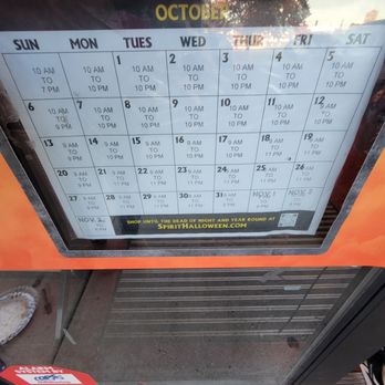 Halloween Express - A printed calendar for October is displayed inside a glass case, showing daily business hours from 10 AM to 10 PM, with some days closing earlier or opening later. The calendar is organized by week, with each day labeled and hours specified. A promotional banner at the bottom advertises Halloween-themed shopping at SpiritHalloween.com. - Costumes Near Me - Normal, Illinois