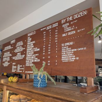 Sunday Bagels - Menu - A wooden menu board lists bagel flavors, spreads, and pricing, with sections for bagels, spreads, and by the dozen options, in a bakery or cafe setting. - Bagels Near Me - Albuquerque, New Mexico