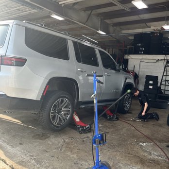 Tirelink - A mechanic is kneeling beside a silver SUV in a garage, using tools for maintenance or repair, with tire racks and equipment in the background. - Tires Near Me - Spring, Texas