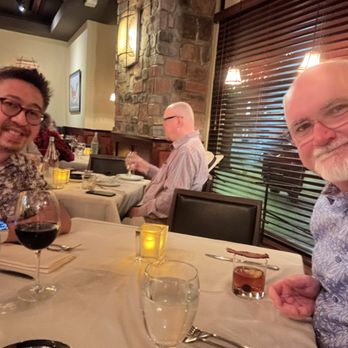Roaring Fork - Two men are seated at a restaurant table with drinks, smiling at the camera. The table has wine, water, and cocktails, with a candle and napkins. The background includes a brick wall, wooden blinds, and warm lighting fixtures, indicating an indoor dining setting. - American Near Me - Austin, Texas