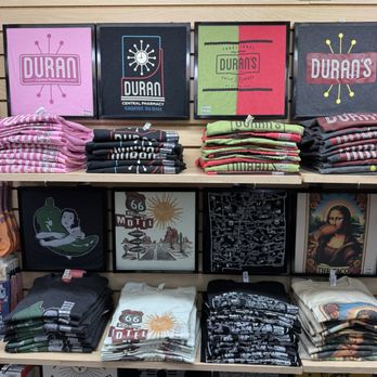 Duran Central Pharmacy - Shelves display folded T-shirts with different graphics and colors, including pink, black, green, and white, at Duran Central Pharmacy in Albuquerque, NM. Above the T-shirts, framed signs with the Duran's logo and branding are visible. The signs include logos and text related to Duran's pharmacy and store. The setup suggests a retail section within the pharmacy that sells apparel and branded merchandise. - Mexican Near Me - Albuquerque, New Mexico