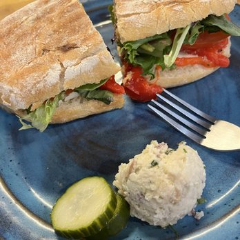 Cloud Forest Cafe - Fresh Mozz Panini - A sandwich made with a crusty baguette contains fresh lettuce, tomato slices, and a red sauce, served on a blue ceramic plate. Alongside the sandwich, there is a pickle slice and a scoop of potato salad with visible herbs and small bits of ingredients. The presentation suggests a casual meal, possibly at a cafe or deli. - Coffee & Tea Near Me - Davis, California