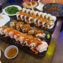 Spicy Tuna - Two black rectangular plates hold assorted sushi rolls with different fillings and toppings, including fish, vegetables, and sauces. A small bowl of soy sauce with mustard seeds is nearby. Additional side dishes include a green salad and a small plate with pickled vegetables. The setting appears to be a restaurant with a wooden table, and a person is seen using chopsticks to pick up sushi. The background includes a blue cup and other dining items.