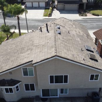 GreenCal Construction - An aerial photograph shows a two-story residential house with a beige stucco exterior and a tiled roof. The roof has multiple vents and skylights. The house is situated in a neighborhood with other homes, a street, and landscaped yards with trees and shrubs. The image captures the layout and surrounding environment of the property. - Roofing Near Me - Bellflower, California