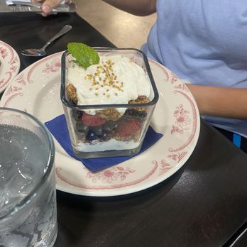 Eggstasy - Yogurt parfait - A layered fruit parfait featuring yogurt, fresh mixed berries, and granola is served in a square glass container. The parfait is topped with a dollop of yogurt, a mint leaf, and sprinkled with granola. It is placed on a decorative plate with a blue napkin underneath. The setting includes a dark table, a glass of water, and a person in a light blue shirt sitting nearby. - Breakfast & Brunch Near Me - Phoenix, Arizona