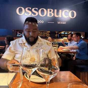 Ossobuco Miami- Steak house - Ossobuco wall - A man sits at a table in Ossobuco Miami, with wine glasses and a menu visible. The restaurant has a modern interior with other diners in the background. - Steakhouses Near Me - Miami, Florida