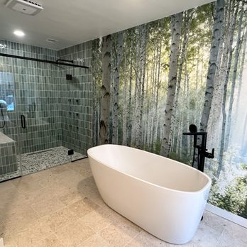 Compass Studio Tile & Flooring - Primary bathroom remodel. - This bathroom has a white, oval-shaped freestanding bathtub positioned against a wall decorated with a forest mural depicting birch trees. To the left, there is a glass-enclosed shower with green tile walls and a pebble tile floor. The bathroom has beige tile flooring and a ceiling with recessed lighting, creating a modern and natural aesthetic. - Carpeting Near Me - Annapolis, Maryland