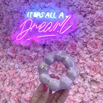 Bober Tea - Taro mochi donut - A hand with pink nail polish holds a lavender glazed donut with a shiny, sparkly surface in front of a wall covered with pink roses. A neon sign in the background displays the phrase 'It was all a dream' in bright pink and blue colors, mounted on the floral wall. The scene has a pastel, dreamy aesthetic. - Donuts Near Me - Carmel, Indiana