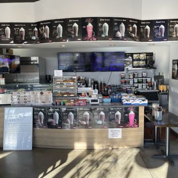 Joker's Koffee - A cafe counter with digital menu boards, beverage displays, and various supplies, with a small table and a fire extinguisher nearby. - Coffee & Tea Near Me - Arcadia, California