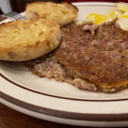 Gritwurst - A breakfast plate includes two slices of toasted bread topped with melted cheese, two fried eggs with runny yolks, and a portion of a cooked meat dish, possibly a type of sausage or steak. The food is arranged on a white plate with a decorative brown rim, placed on a wooden table.