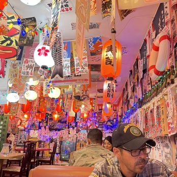 Dragon Sushi - Wonderful decor! - The image shows the interior of a Japanese-themed restaurant with walls and ceiling covered in colorful banners, posters, and flags. Hanging lanterns and string lights illuminate the space, creating a festive atmosphere. Customers are seated at tables, including a man in a plaid shirt and glasses, and a person in military camouflage. The decor includes Japanese characters, images, and traditional elements, contributing to an authentic ambiance. - Sushi Bars Near Me - Albuquerque, New Mexico