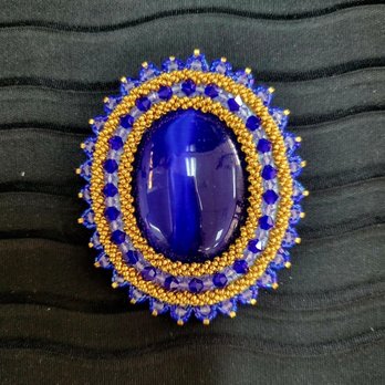 Beaded Iris - A decorative brooch featuring a large, polished blue stone at the center, surrounded by detailed beadwork in blue and gold beads, set against a black fabric background. - Arts & Crafts Near Me - Albuquerque, New Mexico