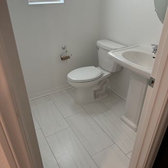 Strusis Tile Installations - A bathroom features a white toilet, a pedestal sink, and a small window with blinds, with white tiled flooring and plain white walls. - Tiling Near Me - Chino, California