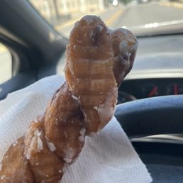 A fried pastry with icing is held on a napkin inside a car, with a view of a street and buildings through the windshield.