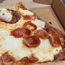 Jumbo Slice Pepperoni Pizza - A close-up image of a pepperoni pizza slice showing melted cheese, crispy crust edges, and pepperoni toppings. The pizza is in a brown cardboard box, with part of the box's edge visible in the background. The image highlights the texture of the cheese and the appearance of the pepperoni slices.