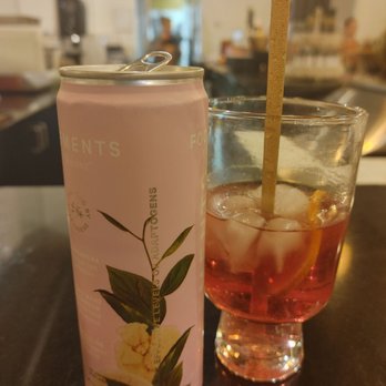 Mila's Mesa - Refreshing and good. - A pink canned beverage with botanical illustrations and text is positioned next to a glass filled with a pink drink, ice cubes, a lemon slice, and a long straw. The setting appears to be a bar or restaurant with a blurred background of the interior, including lighting fixtures and patrons. - Caterers Near Me - Albuquerque, New Mexico