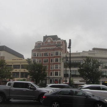 Kenmore Square - A city street scene features several multi-story buildings with varied architectural styles, including a prominent red-brick building with rounded windows. Trees line the sidewalk, and parked cars, including trucks and sedans, are visible along the street. The sky is overcast, creating a gray atmosphere. A McDonald's sign is partially visible on one of the buildings, indicating commercial activity. - Landmarks & Historical Buildings Near Me - Boston, Massachusetts