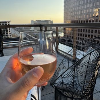 Edge Rooftop - A person is holding a glass of rosé wine on a rooftop terrace with a view of Austin's city skyline, including tall office buildings. The terrace has modern wire chairs and a railing, with the sky showing a sunset or early evening light. The scene suggests a relaxed social setting at an outdoor bar or lounge. - Lounges Near Me - Austin, Texas