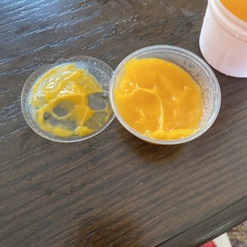 Poor Boy 2 - rubber cheese sauce - Two clear plastic containers hold yellow mango pudding, one with a thicker consistency and the other with a more liquid form, on a wooden table. - Fast Food Near Me - Wheaton, Illinois