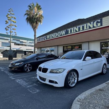 Tint Pros Modesto - Vehicles tinted at pro tint - The image shows two cars parked side by side in front of a window tinting shop. The black car is positioned behind the white car, which is closer to the camera. The shop has a sign indicating window tinting services, and the background includes a tall palm tree and a modern building. The sky is clear and blue, and the parking lot appears clean and well-maintained. - Car Window Tinting Near Me - Modesto, California