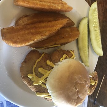 The Bay - Pork tenderloin with wedges - A plate contains a fried fish sandwich with a bun, topped with mustard sauce, and fried fish fillet inside. Accompanying the sandwich are several potato wedges and two slices of cucumber pickles. The plate is placed on a wooden table, with a paper menu partially visible underneath. - American Near Me - Celina, Ohio