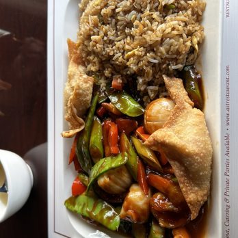 All Seasons Table - Shrimp and scallops with fried rice, crab rangoons. - The image shows a rectangular white plate containing fried rice, stir-fried vegetables including bell peppers, carrots, and onions, and two egg rolls. The fried rice is piled on one side, while the vegetables and egg rolls are arranged on the other side, with a dark sauce underneath the vegetables. The plate is placed on a table with a paper placemat and a small white cup nearby. - Chinese Near Me - Malden, Massachusetts