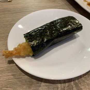Chicago Sushi - Shrimp Tempura hand roll - A shrimp tempura hand roll, wrapped in dark green seaweed, is positioned on a white plate. The roll contains a piece of fried shrimp tail protruding from one end. The plate is on a wooden table, with part of another dish visible in the background. - Japanese Near Me - Des Plaines, Illinois