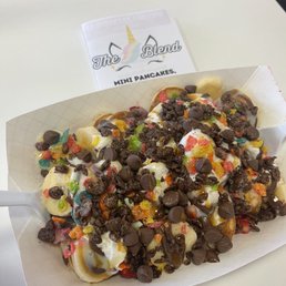 Mini Pancake - Mini pancakes are arranged in a paper tray, topped with chocolate chips, multicolored sprinkles, and syrup. The presentation suggests a dessert or sweet snack, served at a café or casual dining setting. A small sign with the café's name, The Blend, is visible in the background.