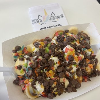 The Blend - Mini Pancakes - Mini pancakes are arranged in a paper tray, topped with chocolate chips, multicolored sprinkles, and syrup. The presentation suggests a dessert or sweet snack, served at a café or casual dining setting. A small sign with the café's name, The Blend, is visible in the background. - Juice Bars & Smoothies Near Me - Albuquerque, New Mexico