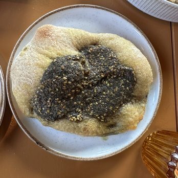 Othello - Za'atar bread - A round piece of bread, likely flatbread, is placed on a white plate. It is topped with a dark greenish za'atar spice mixture, sprinkled with sesame seeds. The bread appears to be baked and dusted with flour or powdered sugar. The dish is part of a meal at a restaurant, with other food items visible nearby. - Seafood Near Me - Jacksonville, Florida