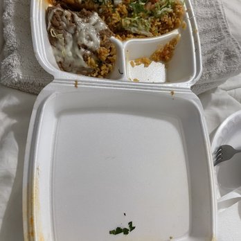 Taco Patio - Food toppled over - A foam takeout container is open, displaying leftover Mexican food including rice, beans, and meat topped with melted cheese. The container has three compartments, with some food remnants and a small piece of green garnish at the bottom. The background includes a towel and a fork on a napkin, indicating a casual dining or takeout setting. - Mexican Near Me - Lockport, Illinois